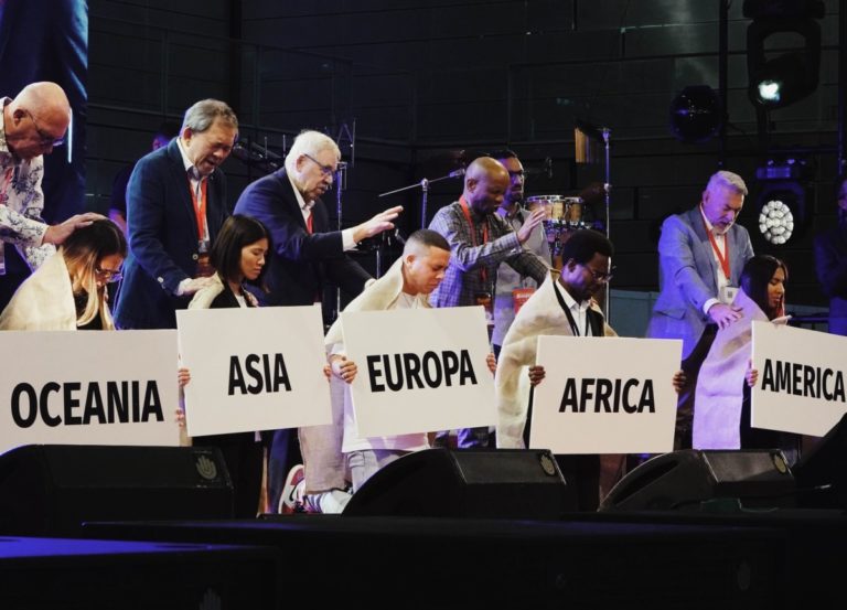 Des personnes prient debout derrière cinq représentants des cinq continents avec une pancarte (Océanie, Asie, Europe, Afrique, Amérique) à genoux