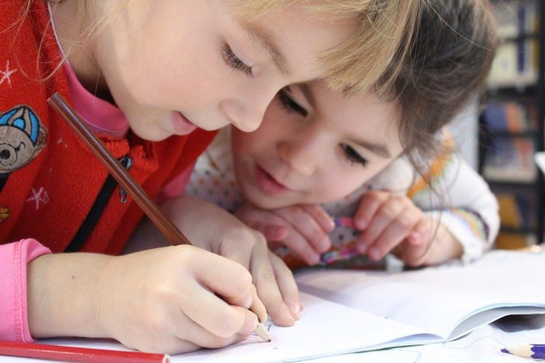 En gros plan, une petite fille blonde écrit sur une feuille. Très près, une autre petite fille penchée observe ce qu'elle fait.