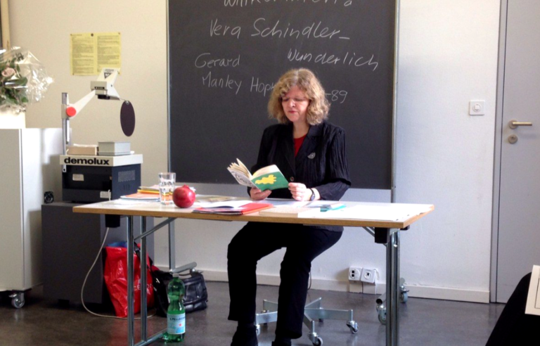 Vera Schindler-Wunderlich, assise derrière un bureau, lit ses poèmes devant une classe de lycéens