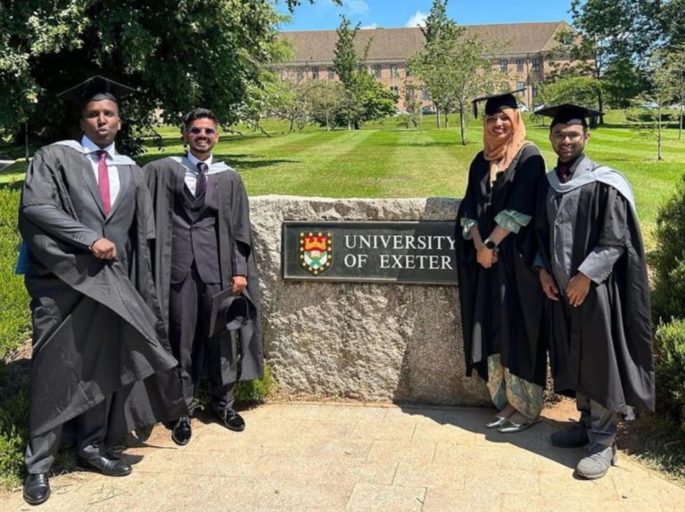 Quatre étudiants diplômés bien habillé autour d'un écriteau "University of Exeter"