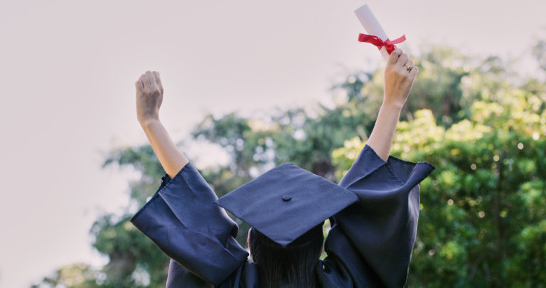 Le haut d'une personne, de dos, habillée avec l'uniforme des diplômés levant les mains vers le ciel, diplôme en main