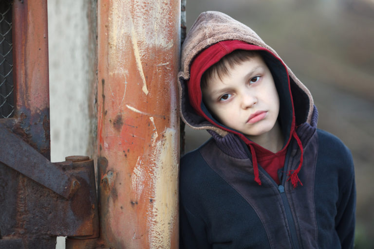 Un enfant, au visage pâle et le regard dans le vide, semble avoir froid dans la rue. Il s'appuie sur un mur