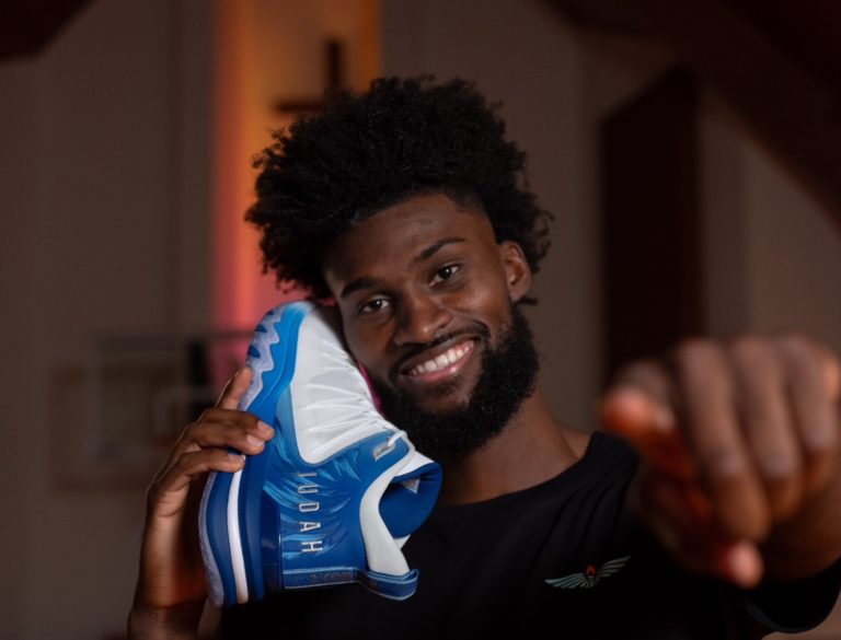 Jonathan Isaac tout sourire, tient sa nouvelle basket avec un verset biblique comme un téléphone. En arrière plan, une croix