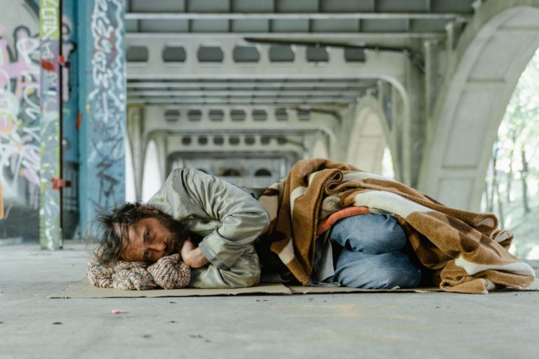 Un homme allongé sous un pont, avec une couverture. En position pour dormir, il a les yeux ouverts et nous fixe.