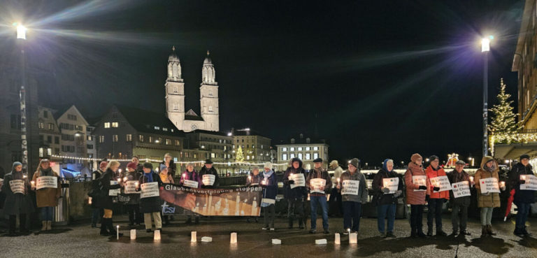 une vingtaine de personnes habillées chaudement sont alignées dans la rue. Elles tiennent des pancartes et des bougies sont posées par terre. Il fait nuit.