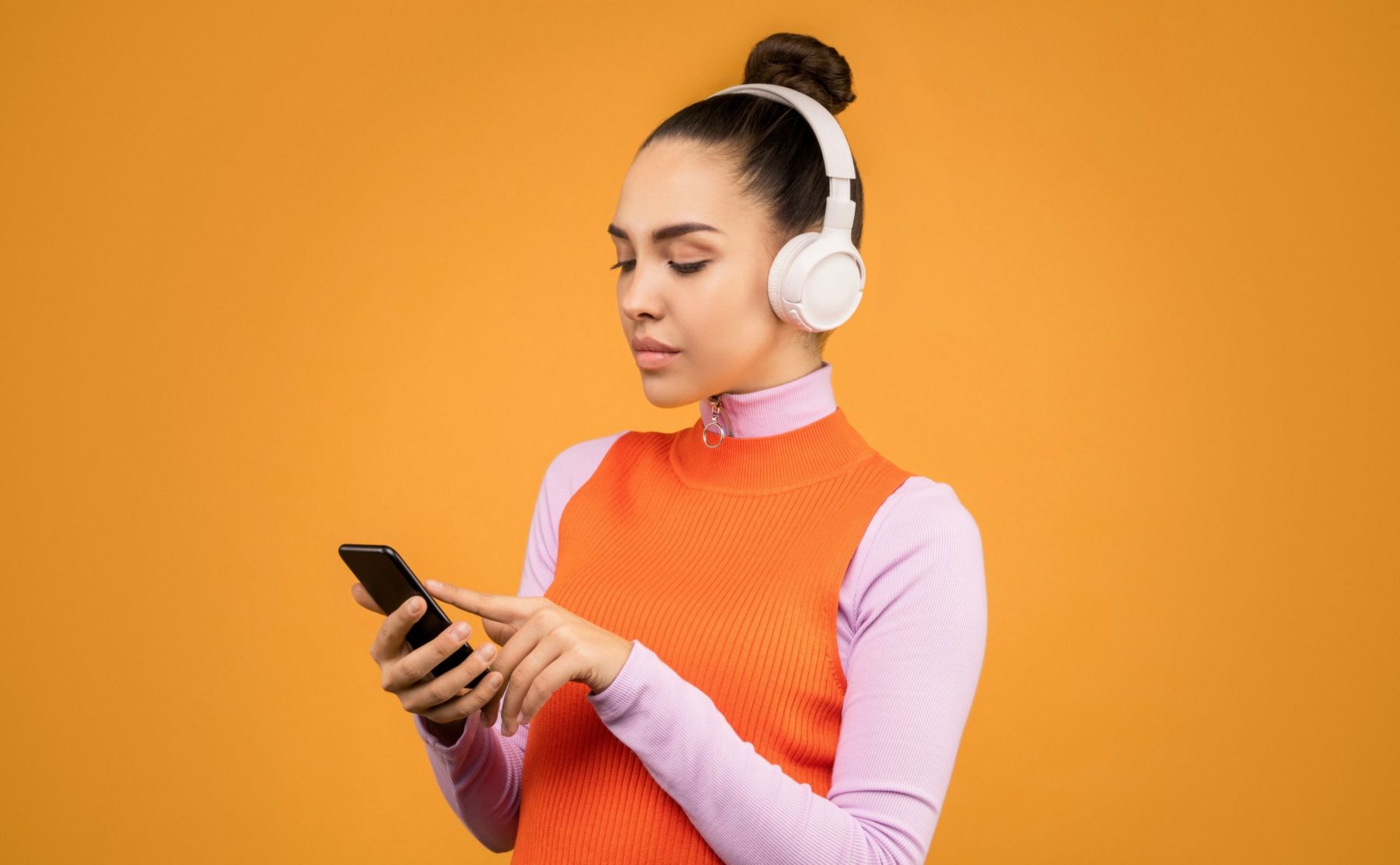 Devant un fond orange, une jeune femme est sur son téléphone. Elle porte un casque pour diffuser de la musique.