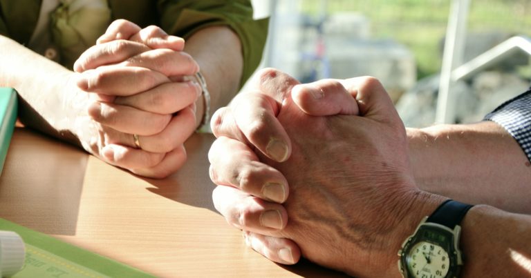 Photo des mains d'un couple d'âge mûr qui prie, les mains jointes posées sur une table. Ils sont en extérieur.