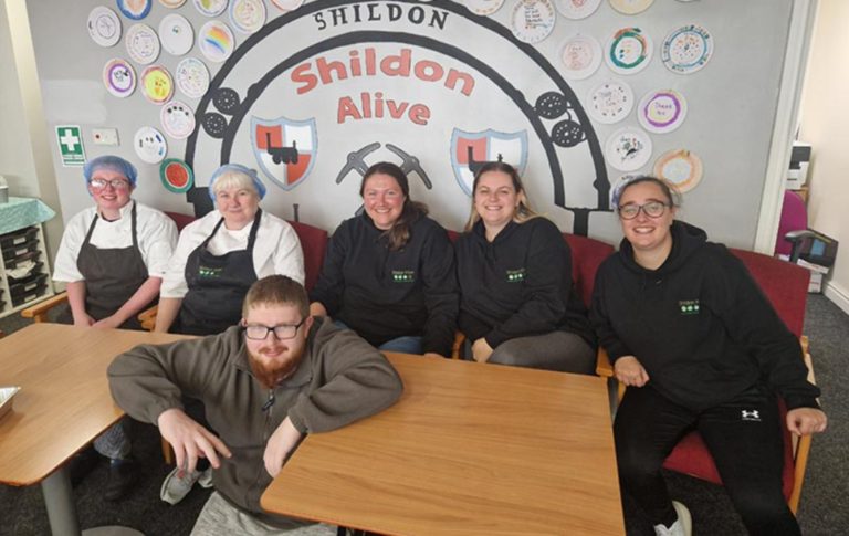 Six bénévoles souriants sont assis, regardant vers l'objectif, derrière deux tables de la cantine du Shildon center. Parmi eux, deux dames sont en habit de cuisinière.