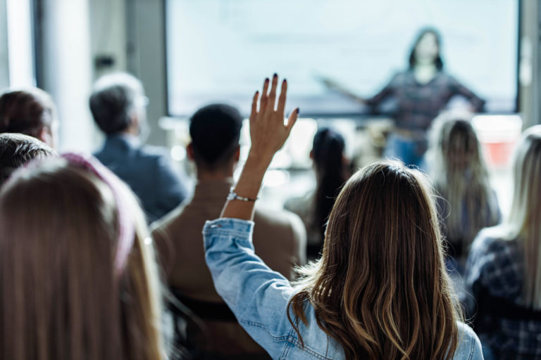 Une conférence avec des personnes assise de dos. Une femme lève la main.