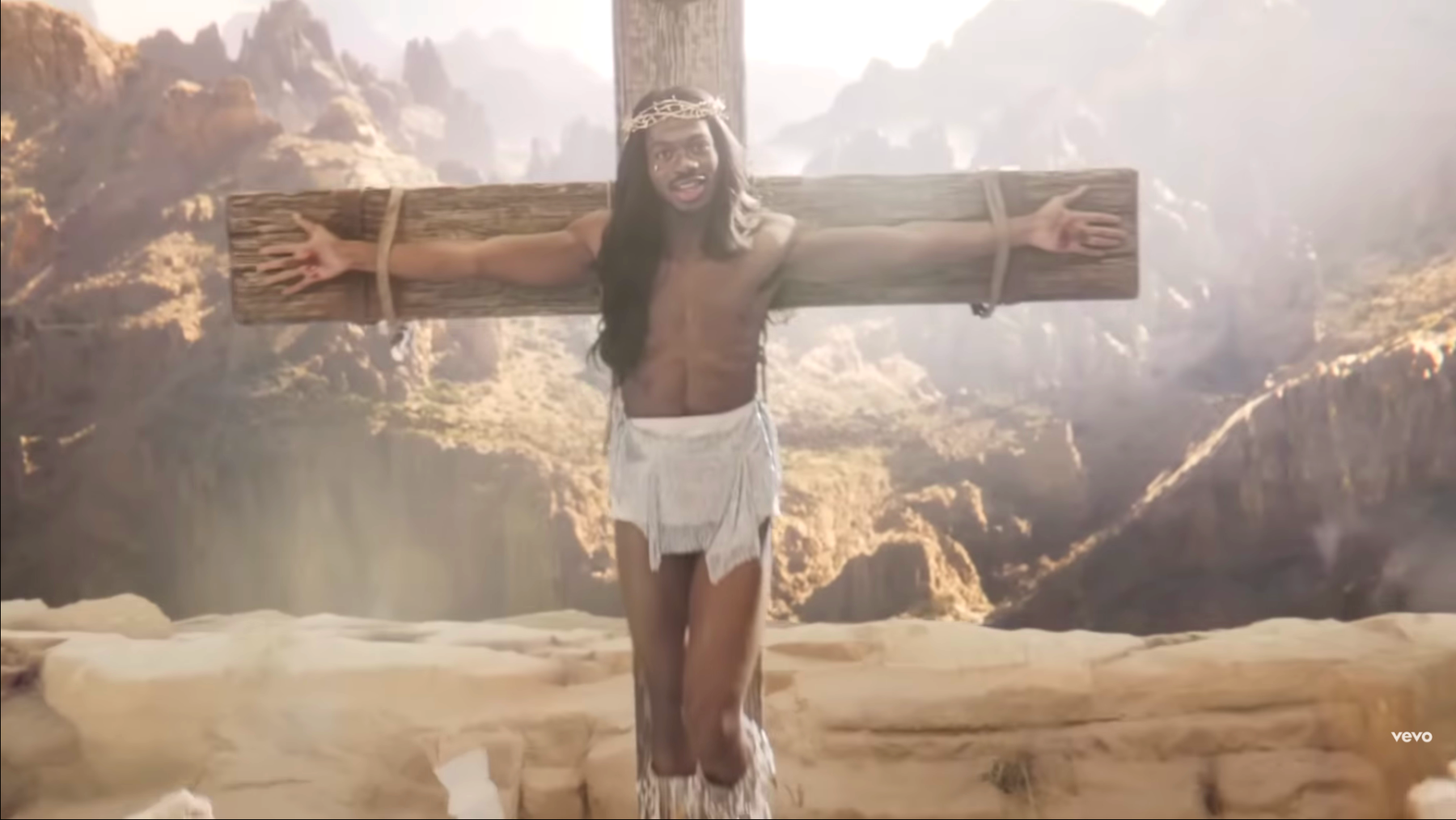 Lil Nas X est attachée à une croix avec les mains percées dans le clip J Christ. Il chante en souriant. Il porte les cheveux longs, une barbe, une couronne d'épines et un pagne blanc.
