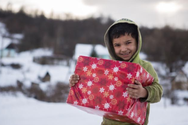 Dans un endroit enneigé, un enfant tient dans ses bras un gros colis enveloppé de papier cadeau