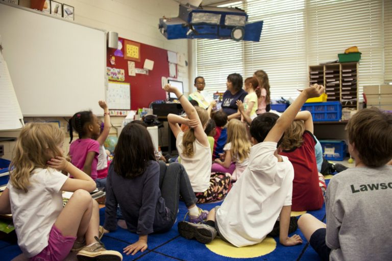 Une classe d'enfants est assise devant son enseignante. Certains lèvent la main.
