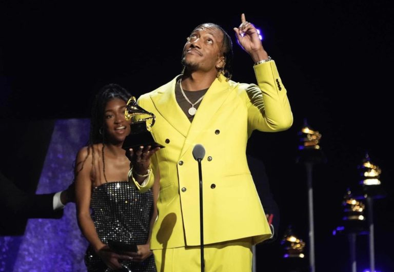 Lecrae avec un costume jaune porte un Grammy Awards dans la main, regarde et lève le doigt vers le ciel pour remercier Dieu