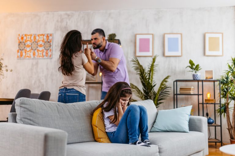 Une femme et un homme se disputent au second plan, tandis qu'une jeune fille assise dans le canapé se bouche les oreilles