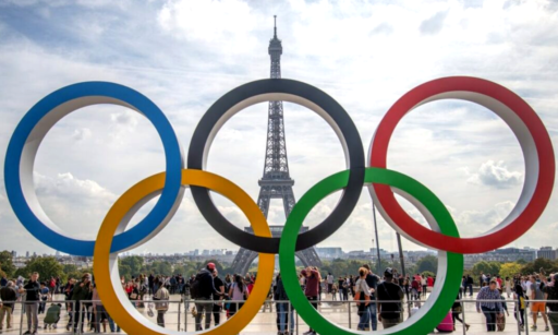 Photo de la tour Eiffel. Au premier plan se trouve une sculpture des 5 anneaux symbolisant les Jeux olympiques