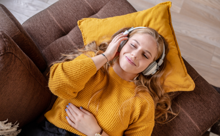 Une jeune femme écoute en souriant, yeux fermés, de la musique avec son casque. Elle est allongée sur son canapé.