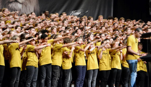 Des dizaine d'enfants chantent sur scène, tous avec le bras droit levé, montrant une direction