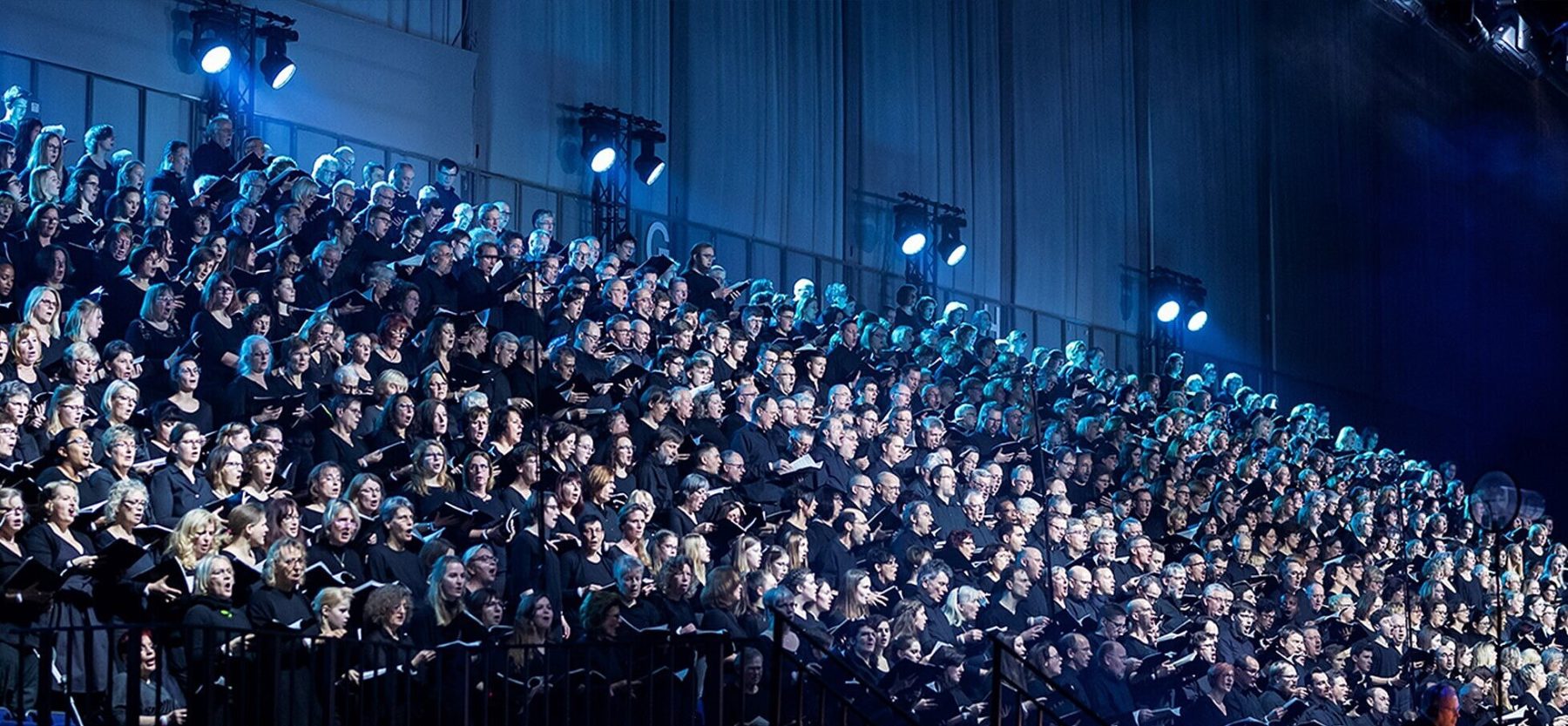 Des centaines de choristes, adultes et enfants, chantent des gradins. Ils ont chacun un livret de chant ouvert dans les mains.