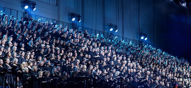 Des centaines de choristes, adultes et enfants, chantent des gradins. Ils ont chacun un livret de chant ouvert dans les mains.