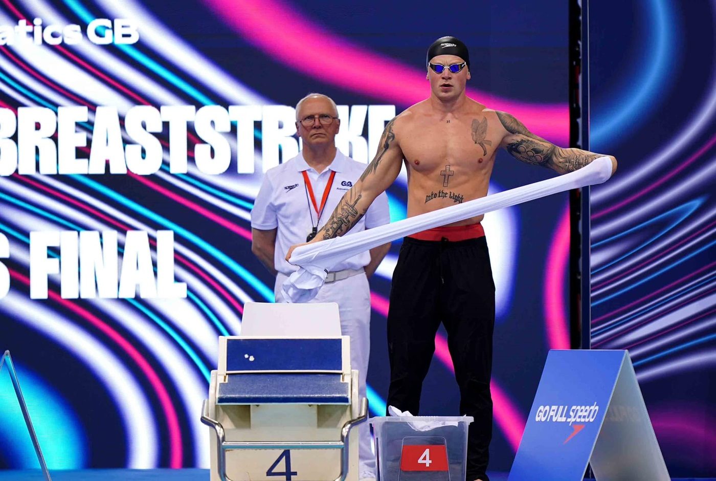 Torse nu, laissant apparaître ses tatouages, Adam Peaty près du plot de départ d'une piscine, il tient une serviette entre ses mains