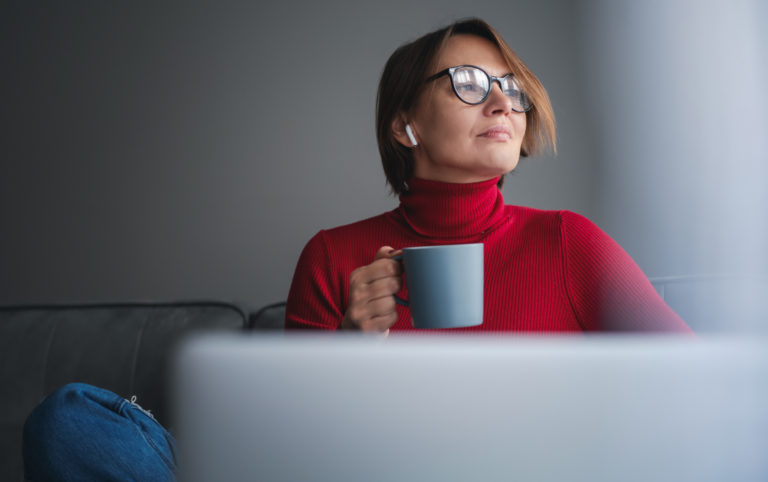 Une dame boit un café devant son PC. Elle a la tête tournée vers la fenêtre