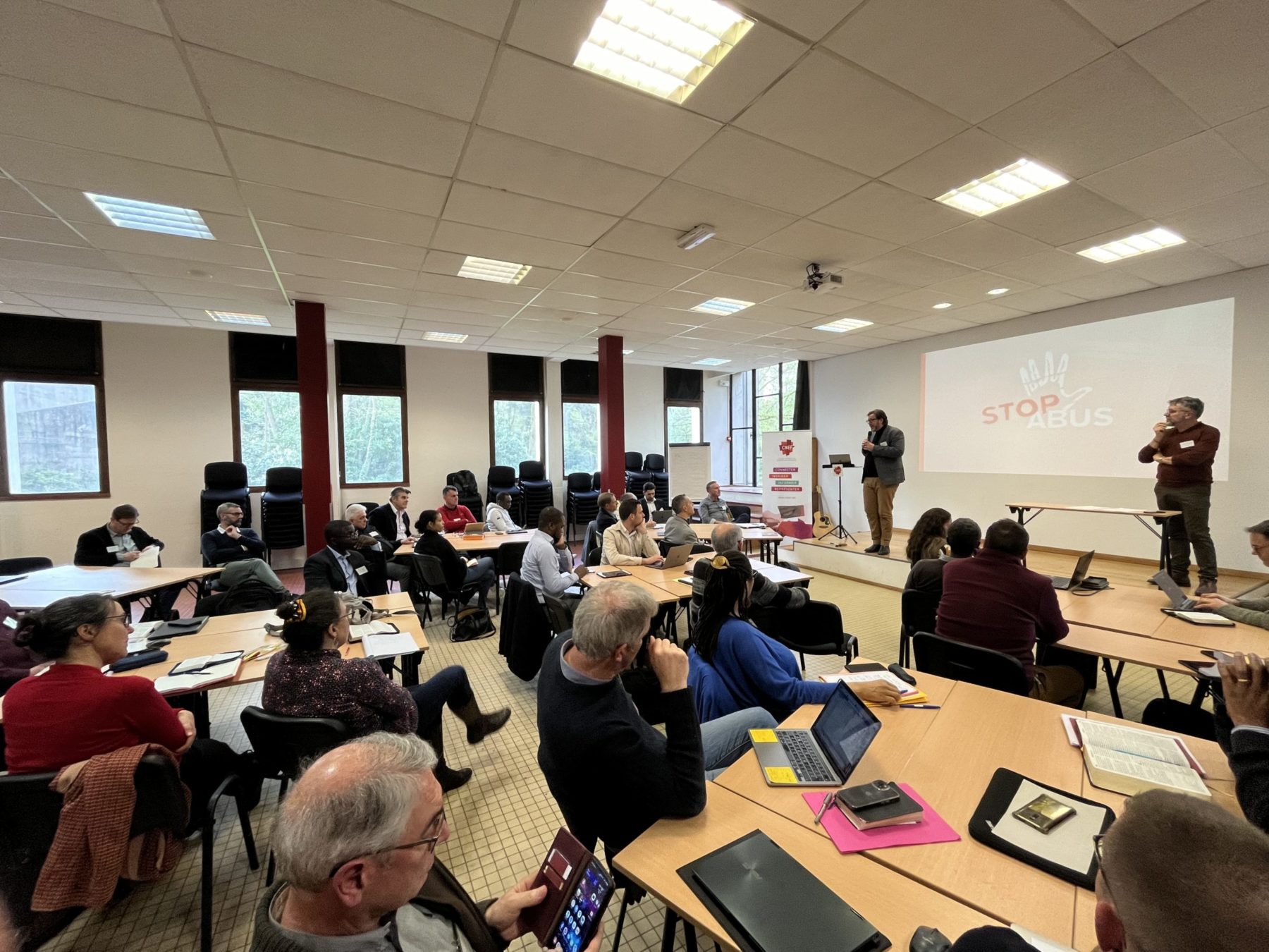 Dans une salle, des personnes sont assises en table ronde écoutent la personne sur l'estrade