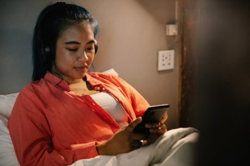 Une jeune femme calée dans son lit, à la lumière d'une lampe, branche ses écouteurs à son smartphone.