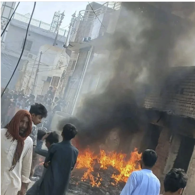 une ruelle est remplie de fumée. des passants regardent un grand feu brûler devant une maison abimée par les flammes