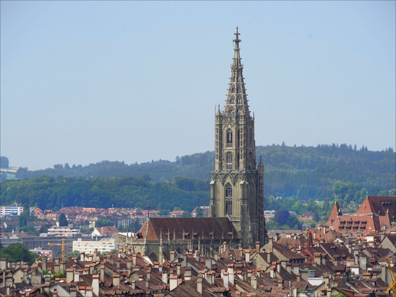 Vue aérienne de la cathédrale de Berne, avec une multitude de toits de maisons au premier plan. Sa flèche est très haute par rapport au reste du bâtiment, qui est moins massif.