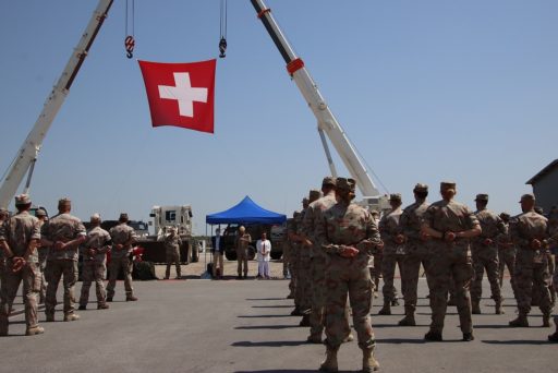 Des militaires debout de dos avec un drapeau de la Suisse suspendu dans les airs grâce à des grues