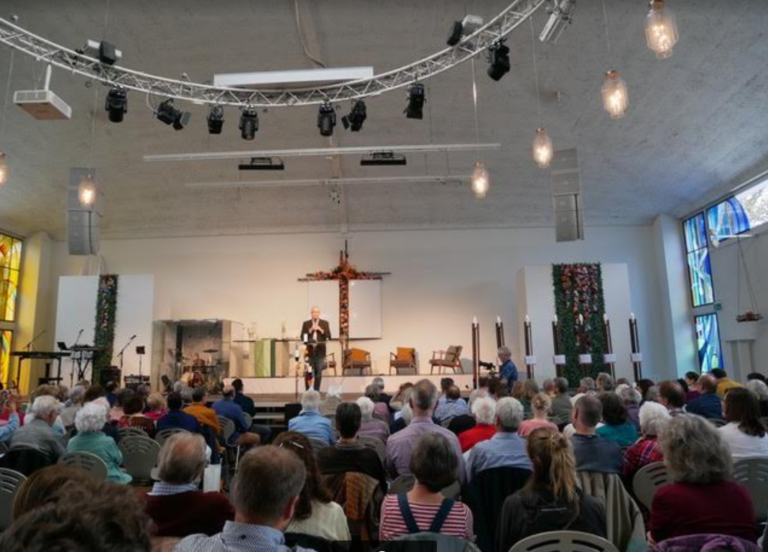 Le directeur de Wycliffe Suisse, Peter Wilburg, s'adresse à un auditoire dans une salle comble. Il est sur une estrade et parle dans un micro.