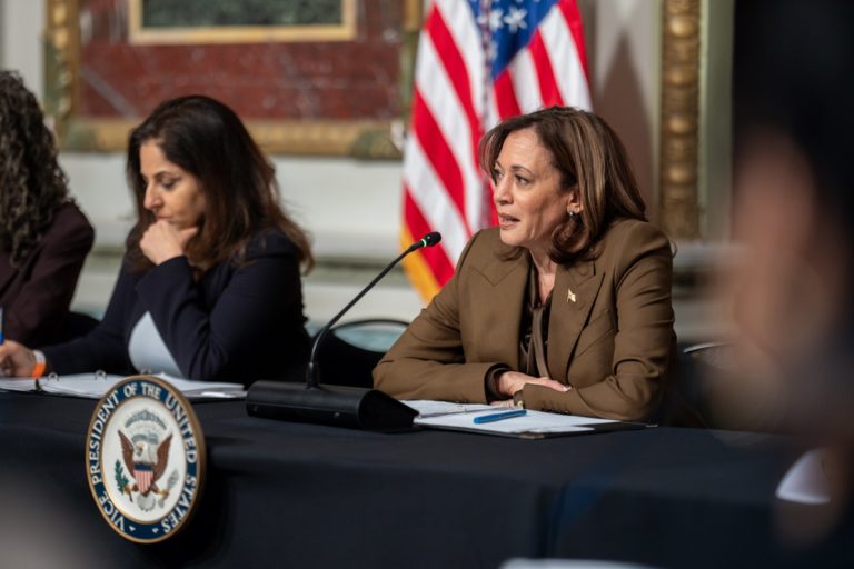 Kamala Harris assise en train de parler dans un micro, le drapeau américain apparaît au second plan