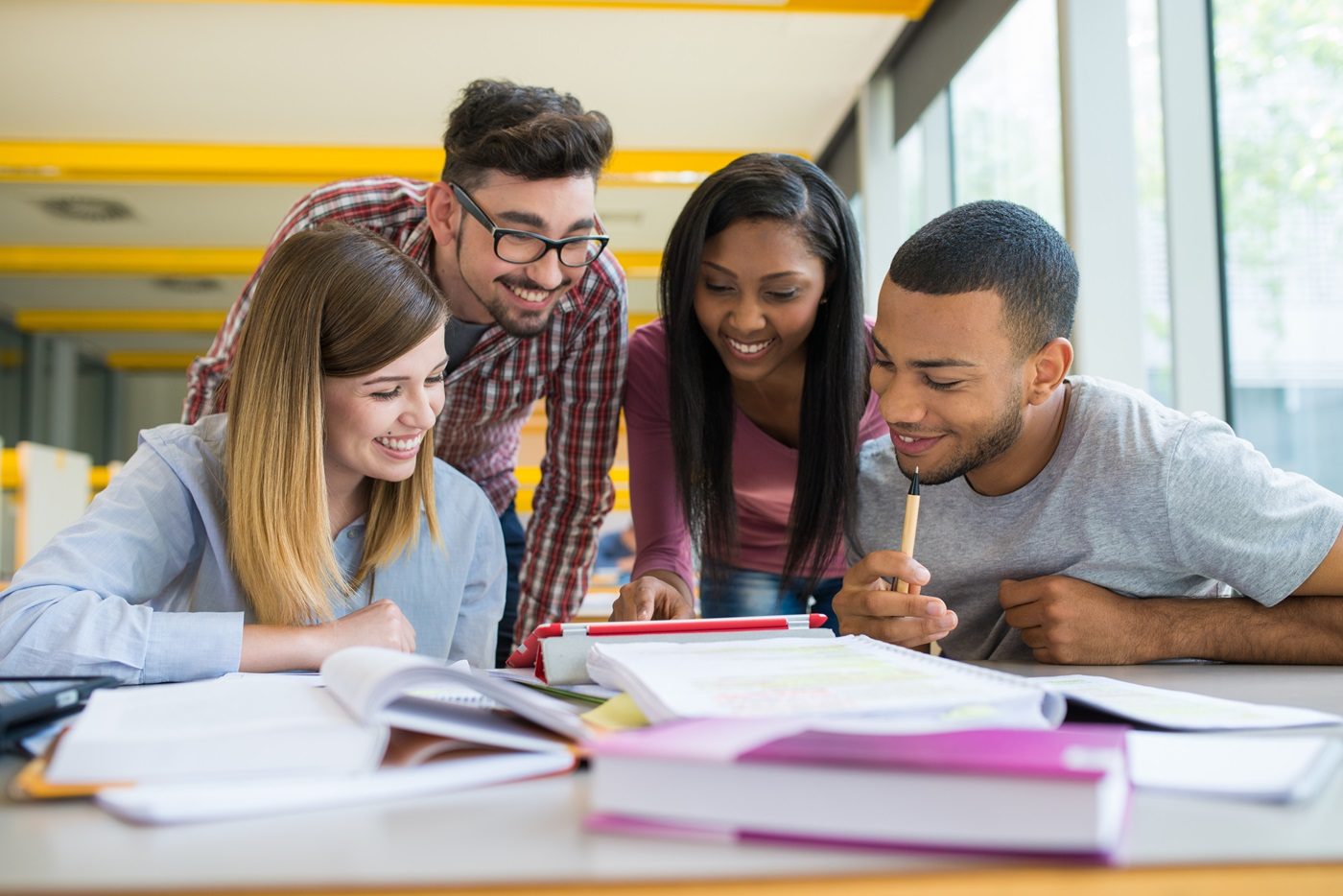 Quatre étudiants avec le sourire en train de regarder une tablette, des cahiers et livres sont posés sur le bureau