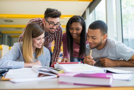 Quatre étudiants avec le sourire en train de regarder une tablette, des cahiers et livres sont posés sur le bureau