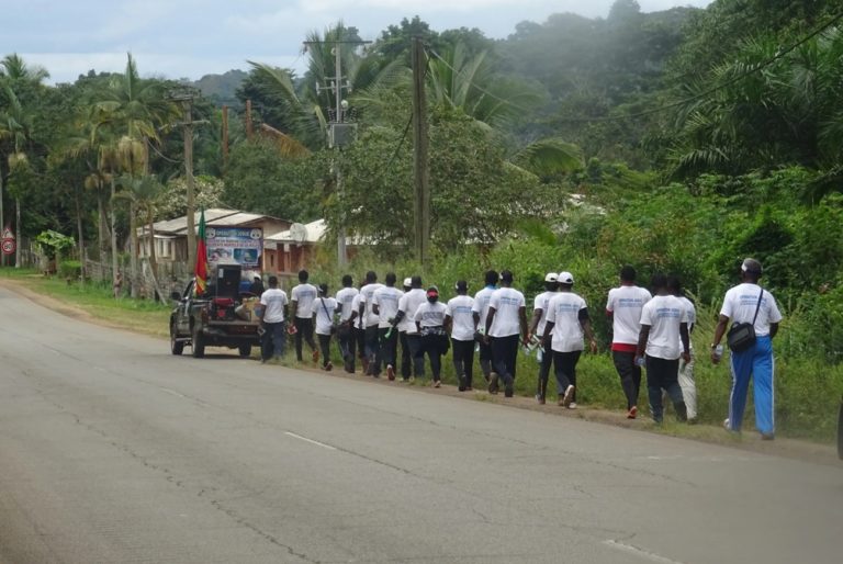 Des hommes et des femmes marchent les uns derrière les autres en suivant un véhicule qui arbore un drapeau du Cameroun