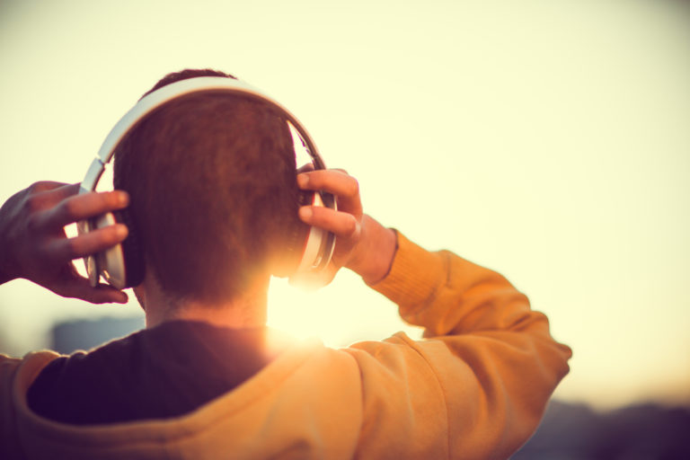 Un jeune homme, deux dos, place un casque sur ses oreilles. Il est en extérieur et le soleil se lève.