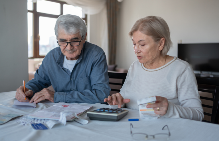 Un couple âgé calcule son budget. L'homme et la femme sont assis à table, avec une calculette et plusieurs documents.