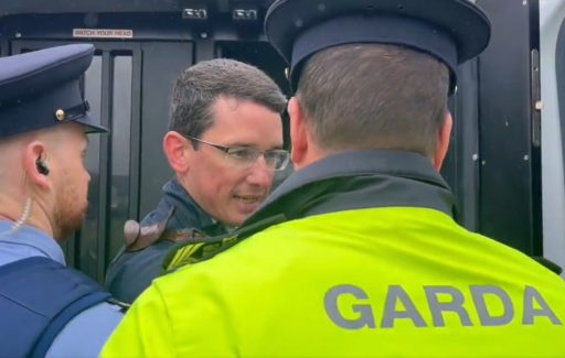 Enoch Burke en train d'être arrêté et mis à l'arrière d'un fourgon de la police irlandaise par deux agents dont l'un avec une veste jaune fluo avec écrit "Garda" dans le dos