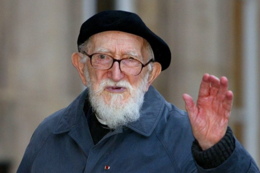 Photo de l'abbé Pierre à un âge avancé. Il lève la main droite, peut-être pour saluer quelqu'un