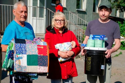 deux hommes et une femme portent dans leurs bras des couvertures, un grand seau fermé et des boîtes de conserve et fourniture d'hygiène