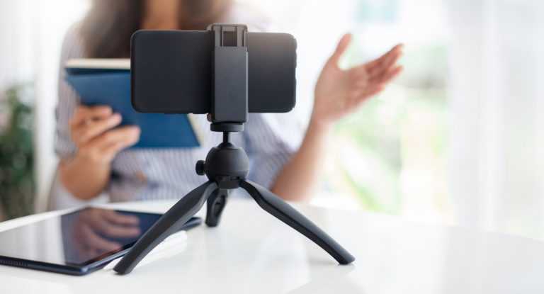 Un smartphone positionné sur un trépied au premier plan. On distingue derrière une femme qui parle en se filmant. Elle tient un livre.