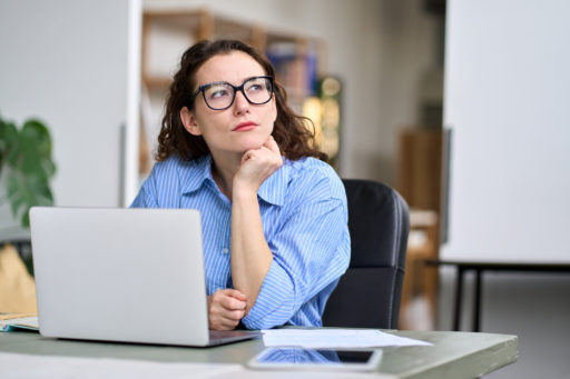 Une jeune femme est assise dans une salle de bibliothèque, son PC posé devant elle. La tête posée sur une main, elle regarde ailleurs.