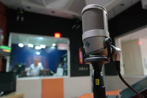 Vue de l'intérieur d'un studio radio avec un microphone sur pied au premier plan et une régie son floutée dans le fond