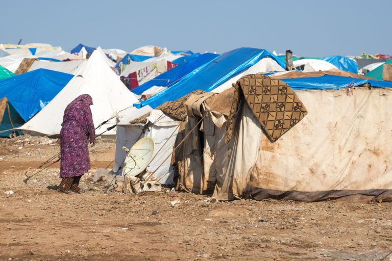 Une dame s'approche d'une tente dans un camp de réfugiés. Des centaines de tentes, en tissu avec des bâches en plastique, sont plantées proches les unes des autres dans un vaste espace de terre, sous le soleil.