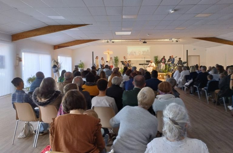 Vue de dos de personnes assises regardant vers une estrade où se trouvent des musiciens, une croix éclairée est attachée au mur