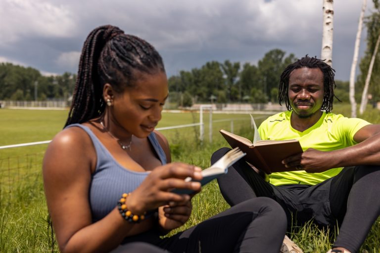 Une femme et un homme lisent chacun leur bible assis sur l'herbe à proximité d'un stade de sport