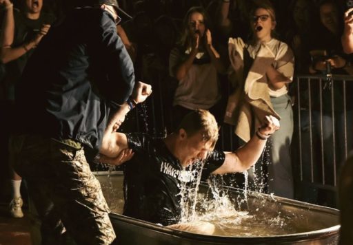 Un étudiant dans un abreuvoir lève les bras après son baptême, il est aidé par un homme pour sortir de l'eau
