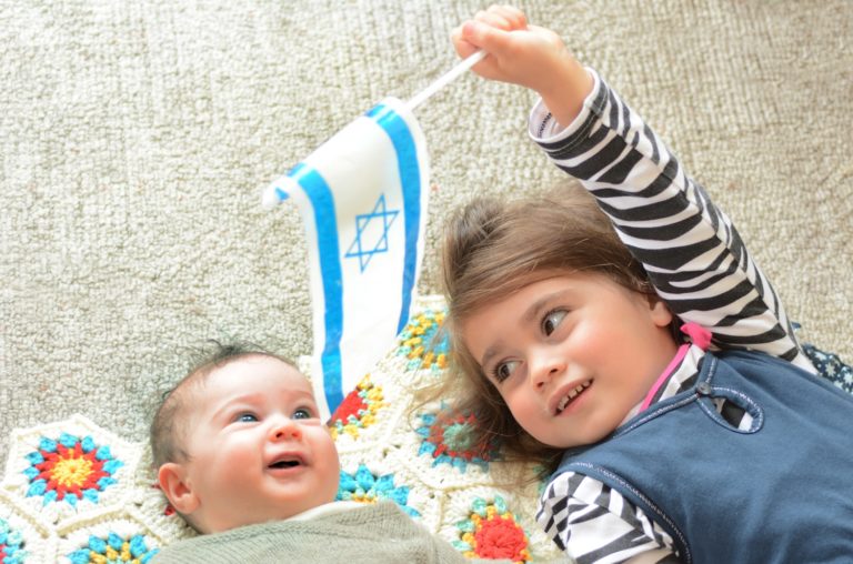 un bébé allongé et sa grande soeur à ses côtés tenant dans sa main un drapeau d'Israël (avec la croix de David)