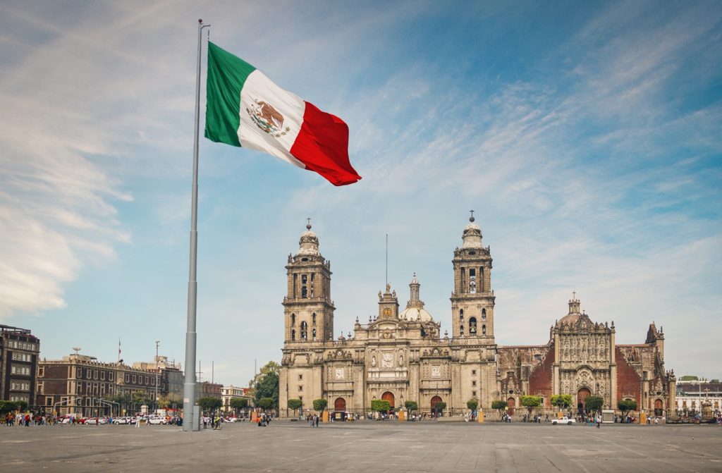 un drapeau mexicain trône sur une place de Mexico, avec au second plan la cathédrale de la ville