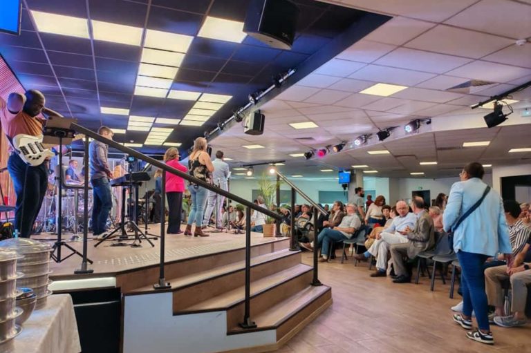 l'intérieur d'une salle de culte avec une estrade sur laquelle sont musiciens et chanteurs, en bas des personnes sont assises